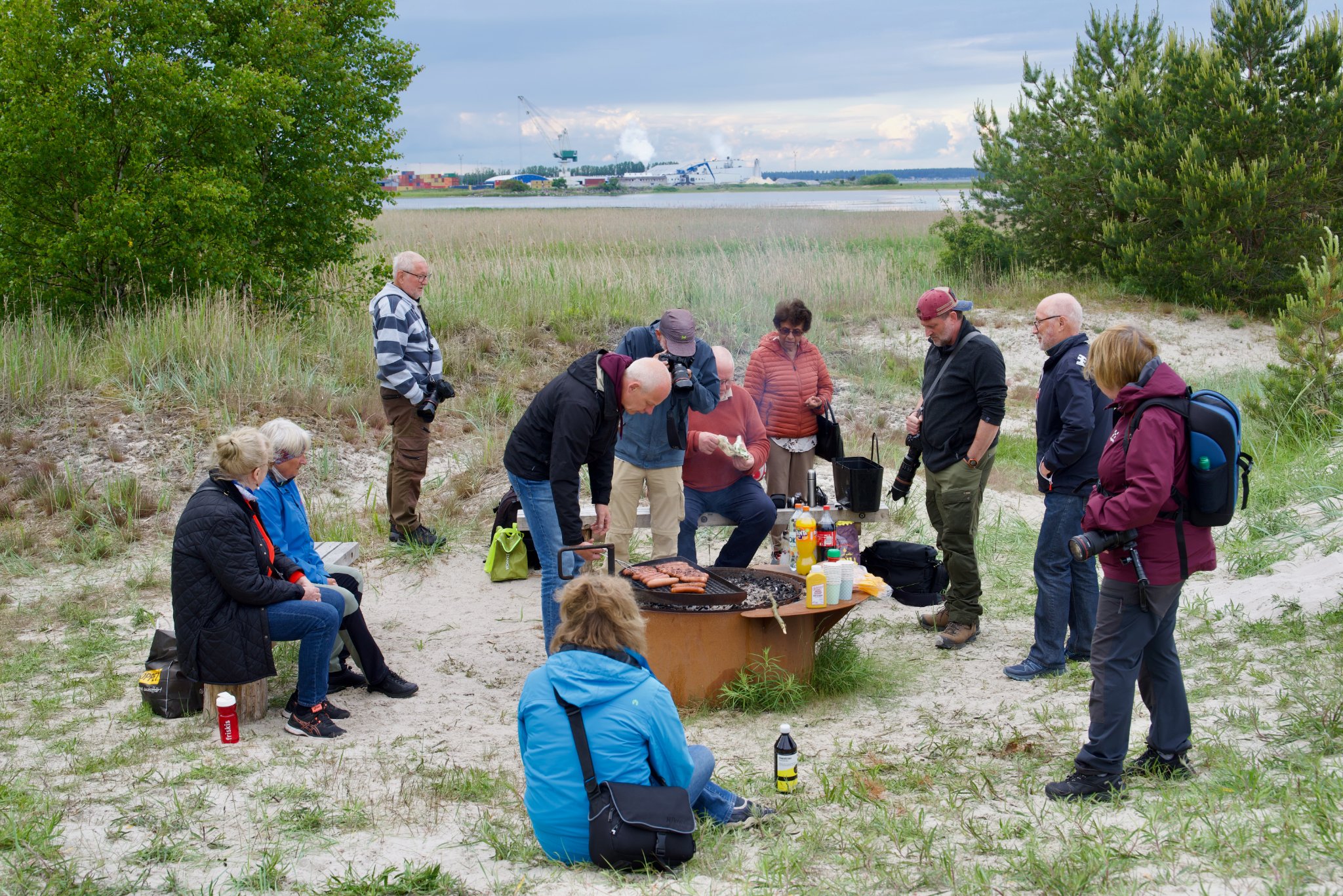 Ett gäng personer befinner sig i naturen samlade kring en eldplats och grillar.